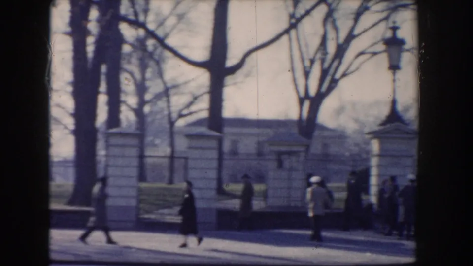 Several pedestrians, including a woman in a dark coat, walk near stone posts and bare trees in a Washington DC street scene from 1960.