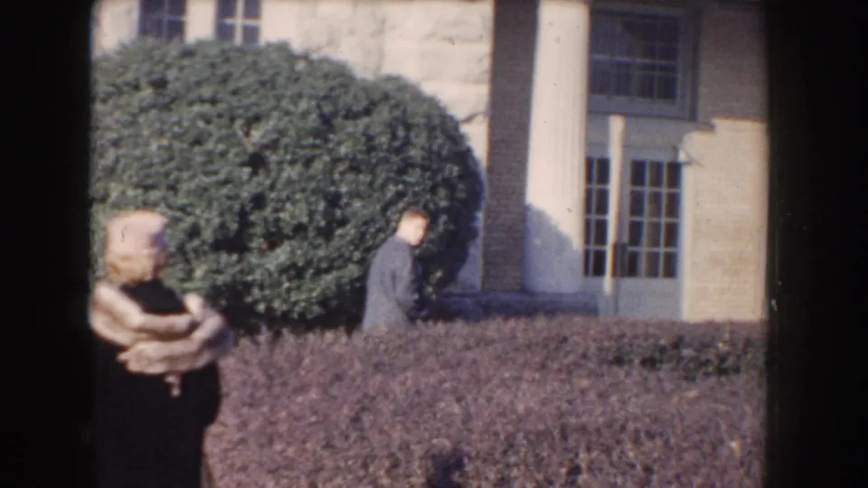 1960 Washington, DC frame with a woman in hat and fur stole near a large shrub as a suited man approaches the columned entrance.