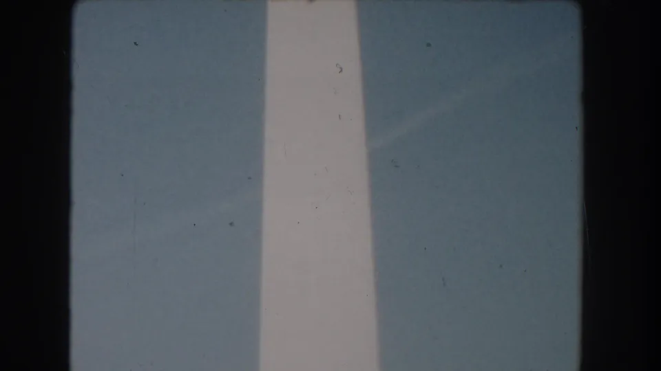 1960s 8mm view of the Washington Monument shaft cropped close against clear blue sky, with visible grain and gate edges.