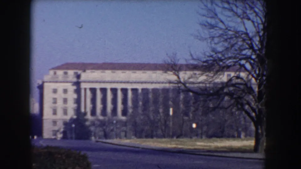 Washington, DC civic architecture with a columned facade, winter trees, lawn, and roadway in soft 1960 8mm footage.