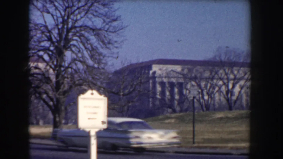 White mid-century sedan passes a roadside sign in Washington, DC, with formal architecture and bare trees behind it.