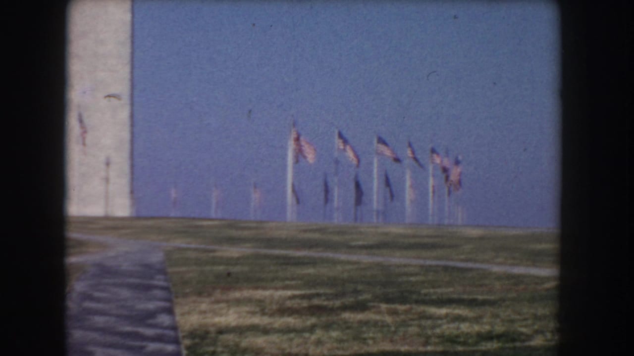 1960 Washington, DC frame with the Washington Monument, American flags, grass, and paved paths under a clear blue sky.
