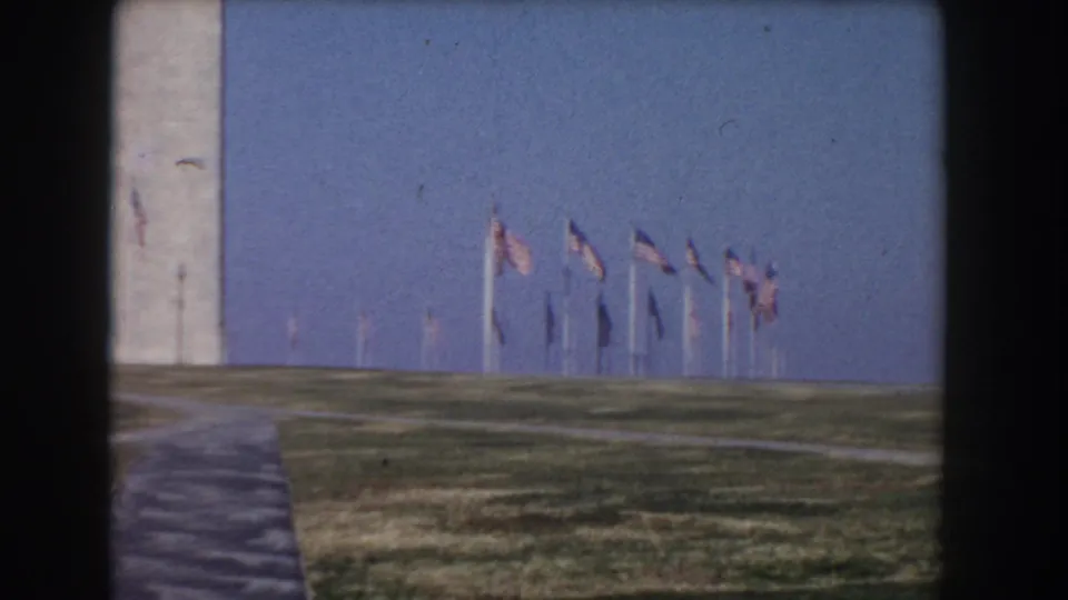 1960 Washington, DC frame with the Washington Monument, American flags, grass, and paved paths under a clear blue sky.