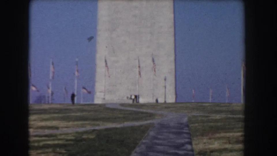 1960 Washington, DC home-movie frame showing the lower Washington Monument, flags, intersecting paths, grass, and distant visitors.