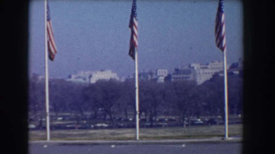 1960 Washington, D.C. home-movie frame with flags in the foreground and distant buildings behind winter-bare trees.
