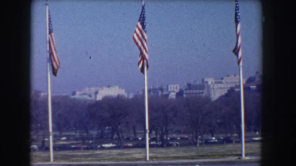 Washington, D.C. 8mm frame showing flags before bare trees, parked cars, and a hazy line of distant buildings.