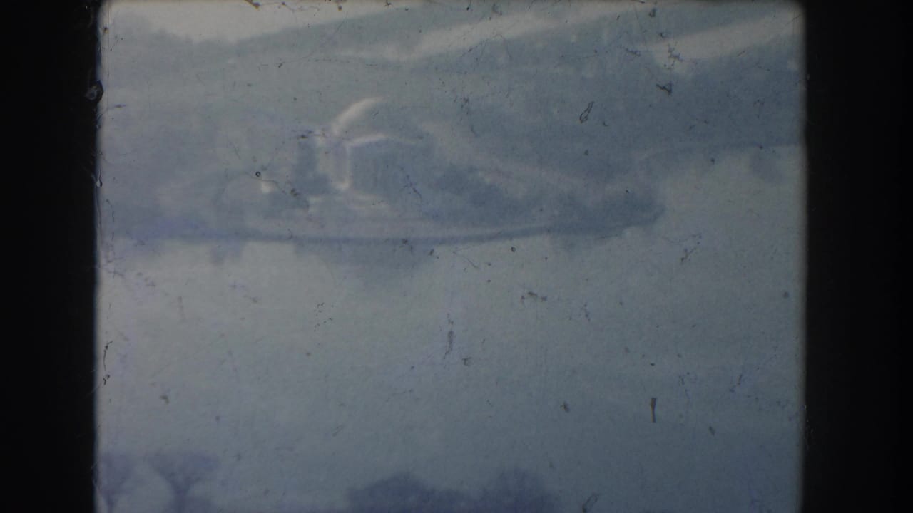 Faded aerial frame with a domed structure near water and visible film scratches.
