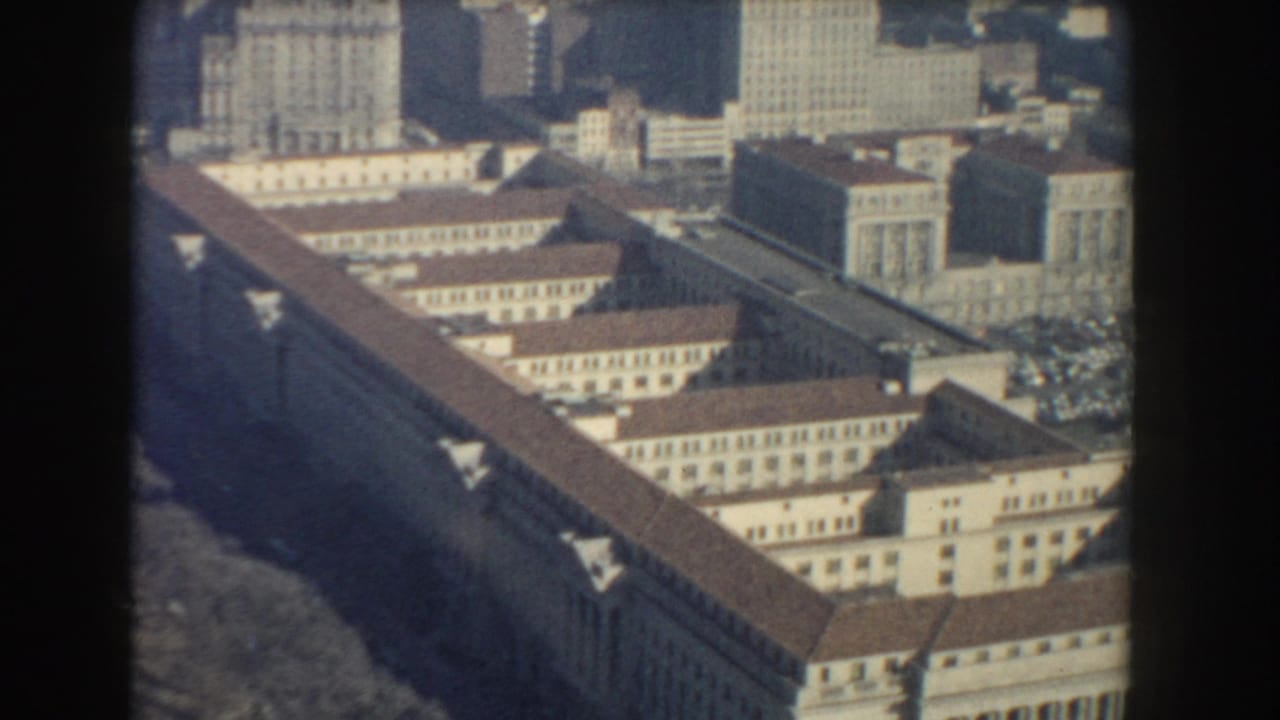 Aerial view of a long pale building with red-brown rooflines and taller city blocks behind it.