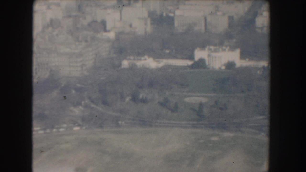 Aerial view over green grounds, roads, and pale buildings in the distance.