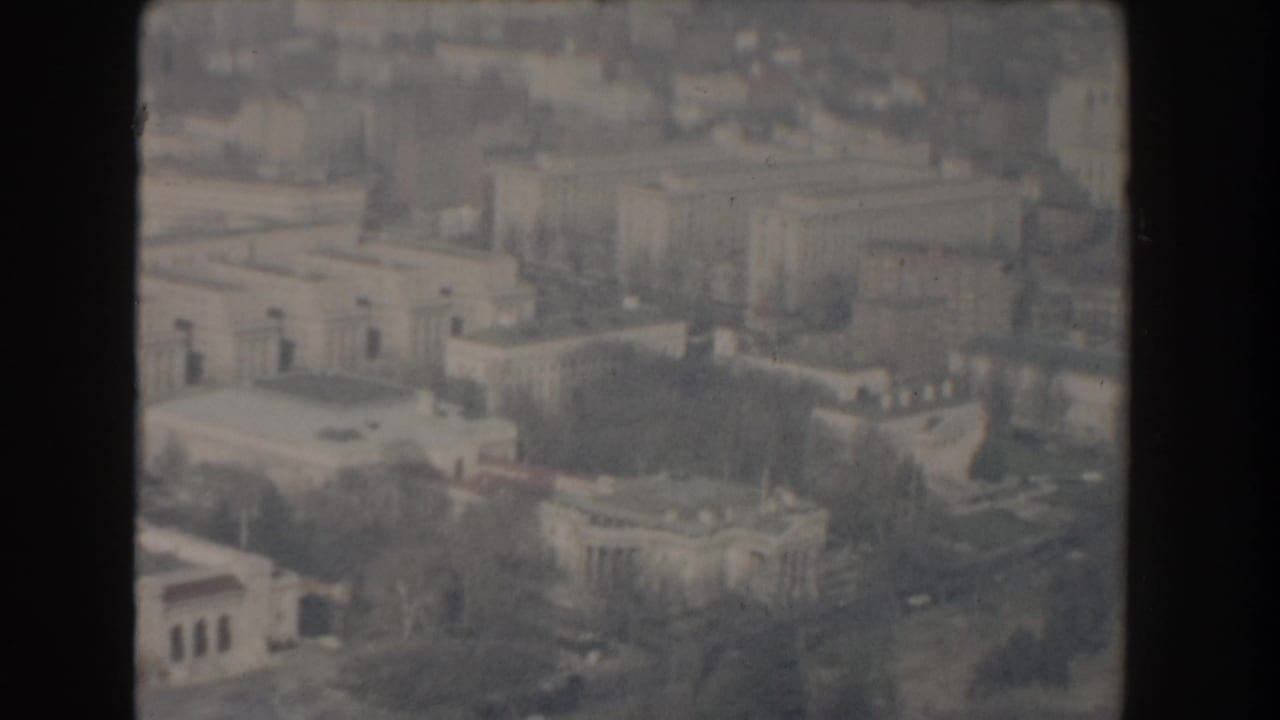 Aerial view of rooftops, streets, and blocky civic buildings in a hazy cityscape.
