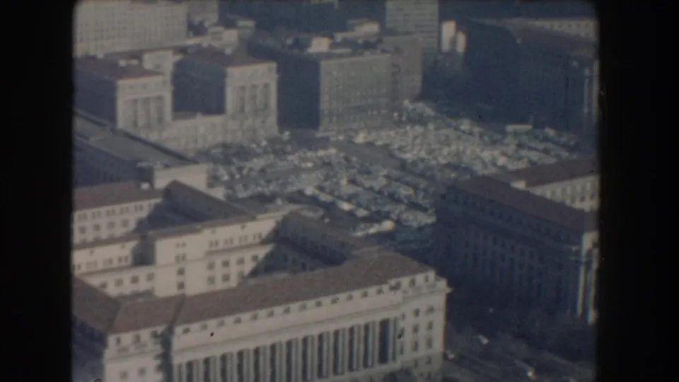 1960 Washington, D.C. aerial frame with classical government-style buildings, office blocks, and a large lot filled with cars.