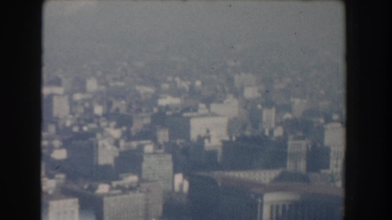 Soft aerial cityscape with dense buildings and a large domed or rounded roof form.