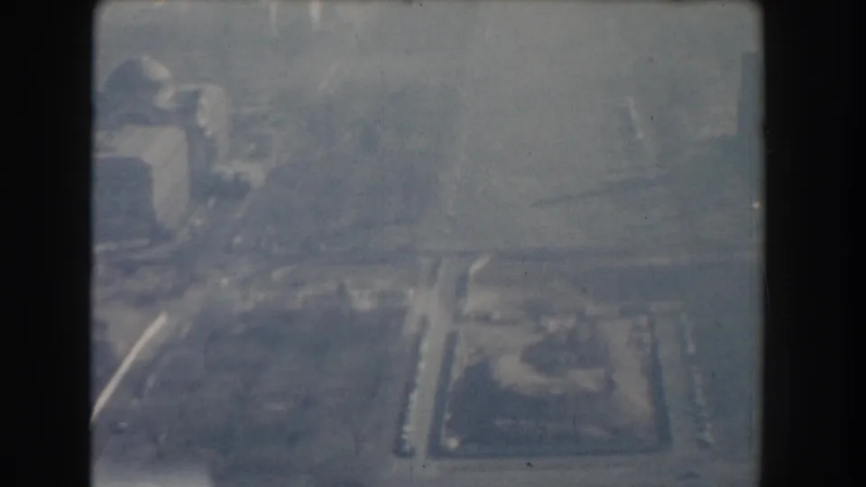 Washington, DC 1960 aerial 8mm frame showing soft-focused buildings, streets, and open ground under blue-gray haze.