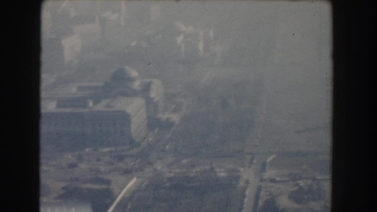 Soft aerial view of buildings, roads, and a bridge approach near water.