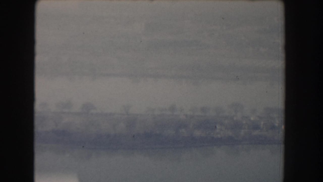 1960 home-movie frame with muted blue-gray water and a faint urban shoreline, possibly from an elevated or moving vantage point.