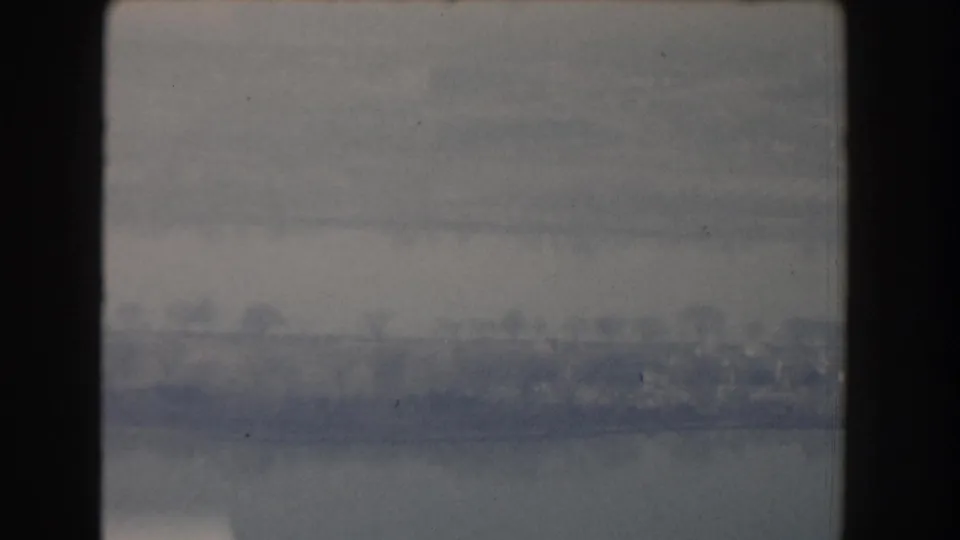 1960 home-movie frame with muted blue-gray water and a faint urban shoreline, possibly from an elevated or moving vantage point.