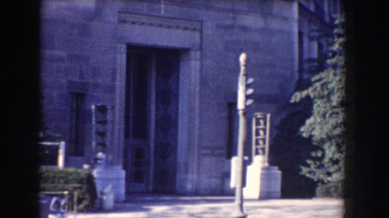 Stone building entrance with sidewalk, post, shrubs, and dark film edge.