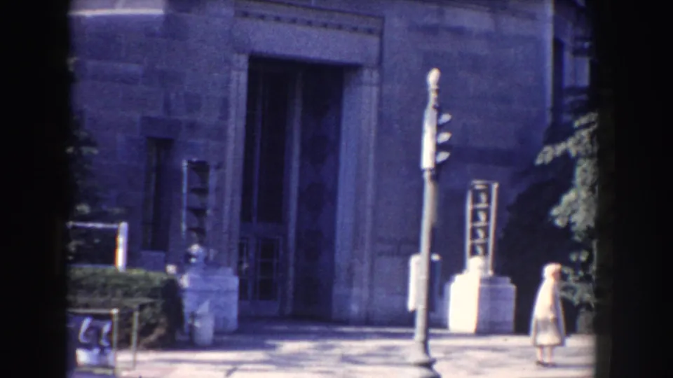 Washington, DC government-building exterior with stone facade, post, shrubs, and a small figure at the right edge.