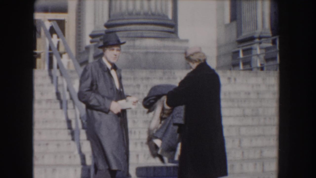 Man in dark suit and hat stands on stone steps beside a woman holding a coat.