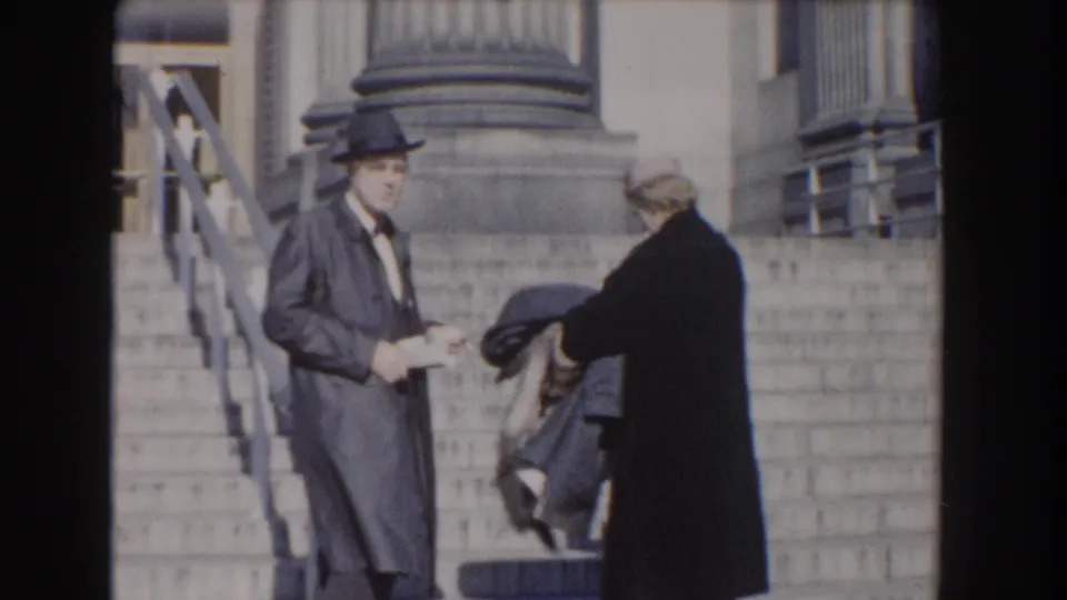 In Washington, DC home-movie footage from 1960, a suited man in a hat stands on broad steps near a woman in a dark coat.