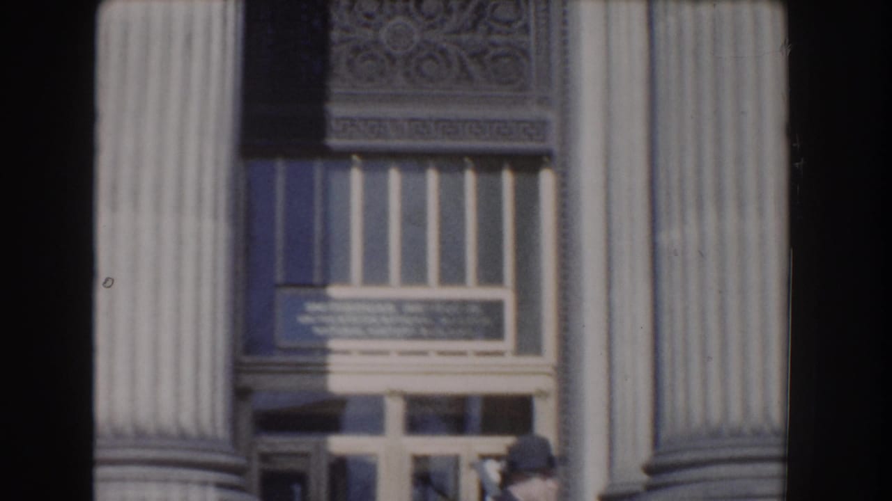 A Washington, DC building entrance appears between tall columns, with signage above the doors and a person partly visible below.
