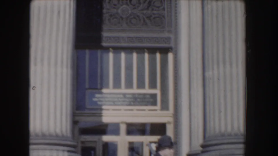 A Washington, DC building entrance appears between tall columns, with signage above the doors and a person partly visible below.