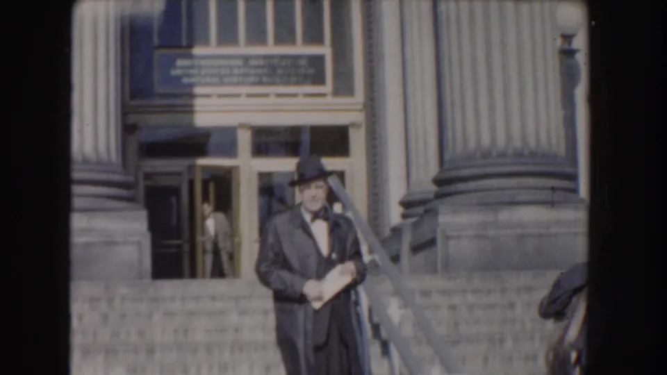 The suited man stands on the steps before a large columned entrance in Washington, DC, with doors and signage visible behind him.