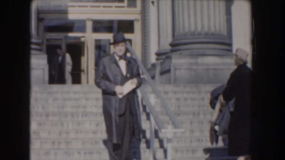 The suited man remains on the Washington, DC building steps as a woman in a dark coat is partly visible near the edge of the frame.