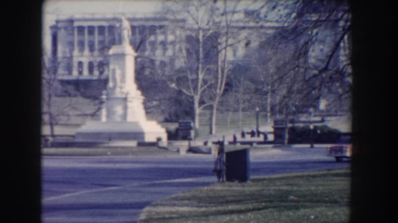 White stone monument in a park with bare trees and large buildings beyond.