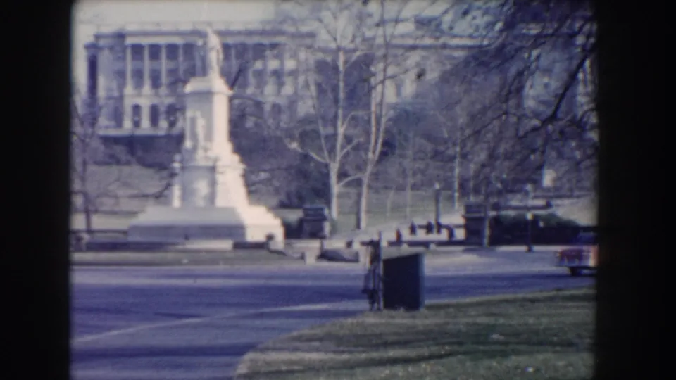 Washington, DC 1960 home-movie frame showing a white stone monument, winter trees, road edge, and civic buildings in the background.