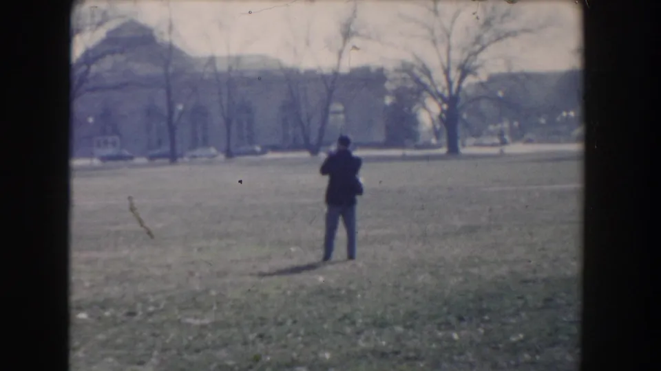 Washington, DC 1960 home-movie frame showing a man in a dark coat standing on grass with a camera near trees and distant buildings.
