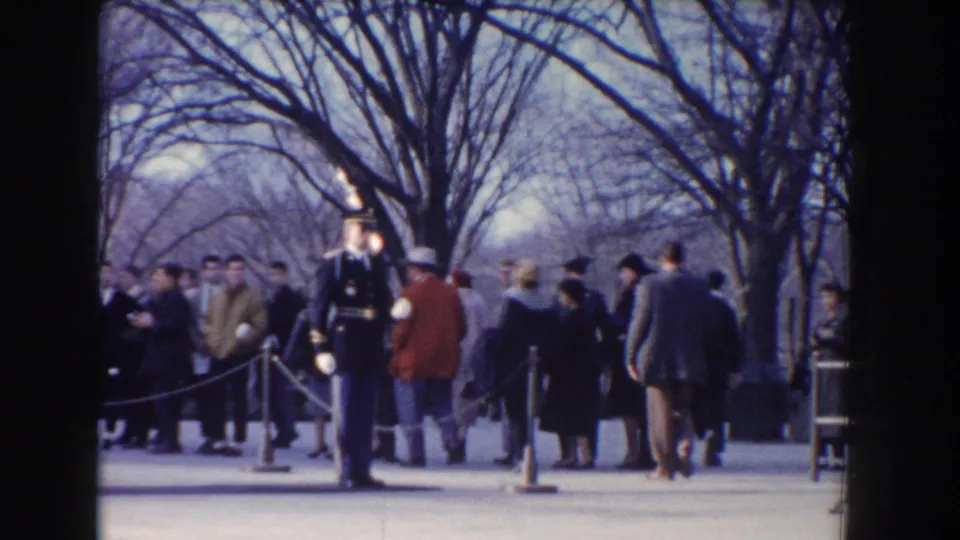 Uniformed ceremonial guard with rifle stands outside in Washington, DC while winter-dressed visitors wait behind ropes.
