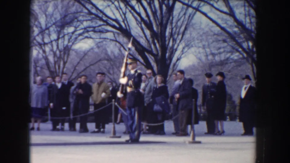 Formal guard with rifle stands before a roped crowd of Washington, DC visitors in winter coats beneath leafless trees.