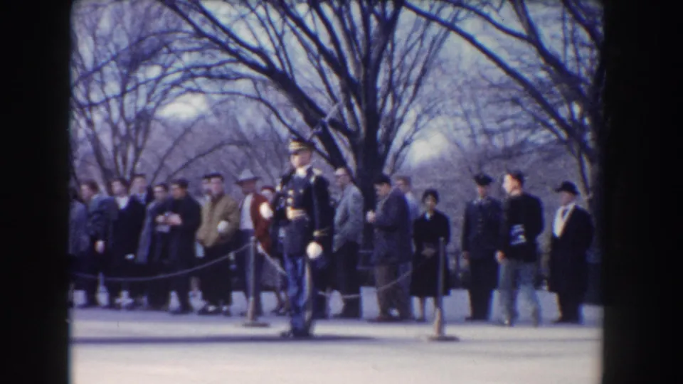 Dress-uniform guard in Washington, DC, with a group of mid-century visitors standing behind a rope barrier under leafless trees.