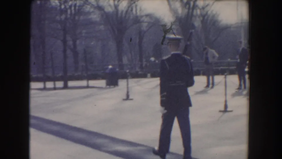Home-movie frame of the ceremonial guard from behind, with rope stanchions, bare trees, and distant visitors in the outdoor public setting.