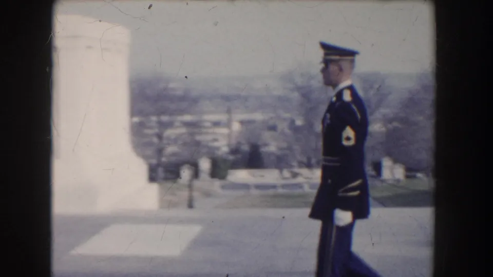 A uniformed guard walks beside a white monument form with a soft distant Washington, DC view in the background.