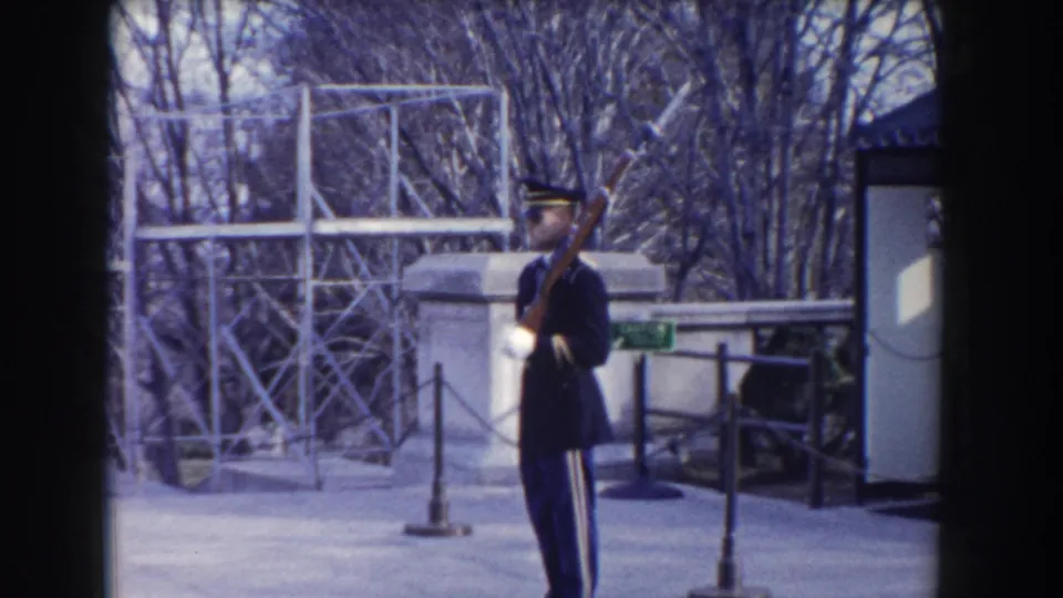 1960 Washington, DC home-movie frame showing a uniformed guard with rifle near rope stanchions and bare trees.