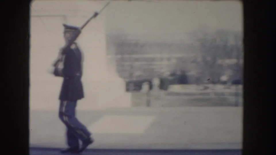 Side view from 1960 Washington, DC footage showing a uniformed guard marching with a rifle, softened by motion blur.