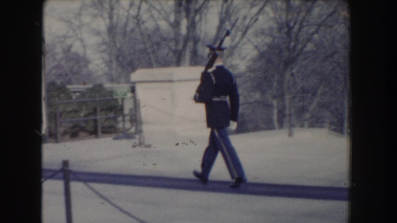 A uniformed guard walks with a rifle on his shoulder beside a low barrier.