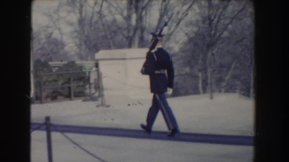 A military guard in dark dress uniform carries a ceremonial rifle while walking through an outdoor Washington, DC setting.