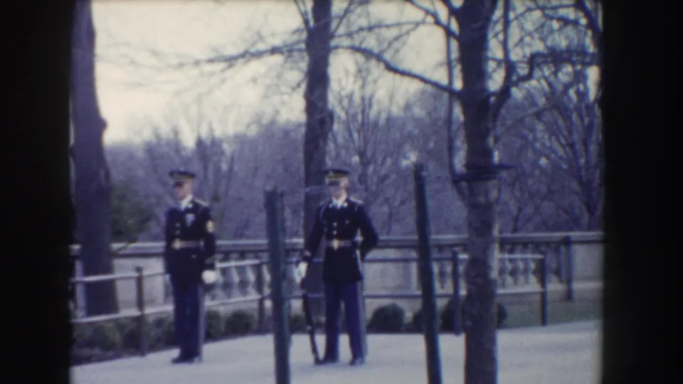 In 1960 Washington, DC footage, two military guards stand separated on a paved walk near leafless trees and stonework.