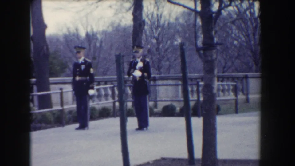 Washington, DC 1960 frame with two Marines in dark dress uniforms and white gloves standing outdoors near trees and a railing.