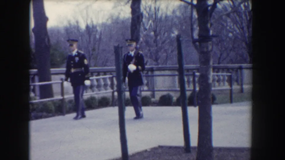 Washington, DC 1960 frame of two Marines in dress uniforms marching in step beside a stone balustrade and bare trees.