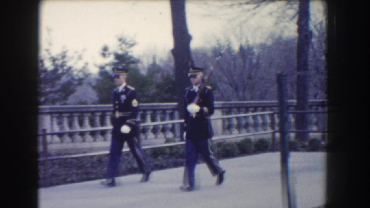 Two military guards in dark uniforms march together in Washington, DC, with rifles and white gloves visible.
