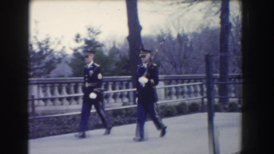 Two military guards in dark uniforms march together in Washington, DC, with rifles and white gloves visible.