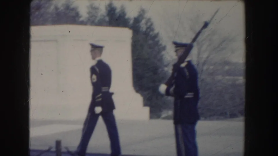 A ceremonial guard stands with a rifle as another uniformed soldier marches past on monument grounds in 1960 Washington, DC footage.