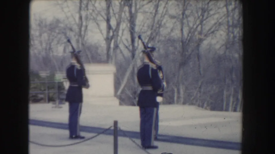 1960 Washington, DC home-movie frame showing two uniformed soldiers with rifles held upright near a low wall and bare trees.