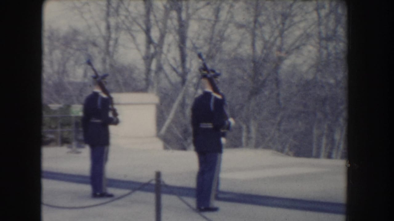 Two uniformed guards stand outdoors near a stone monument and bare trees.
