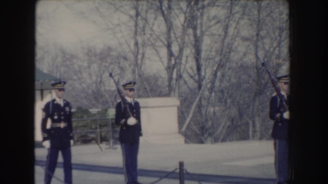 Uniformed guards stand at attention outdoors, one holding a rifle upright.