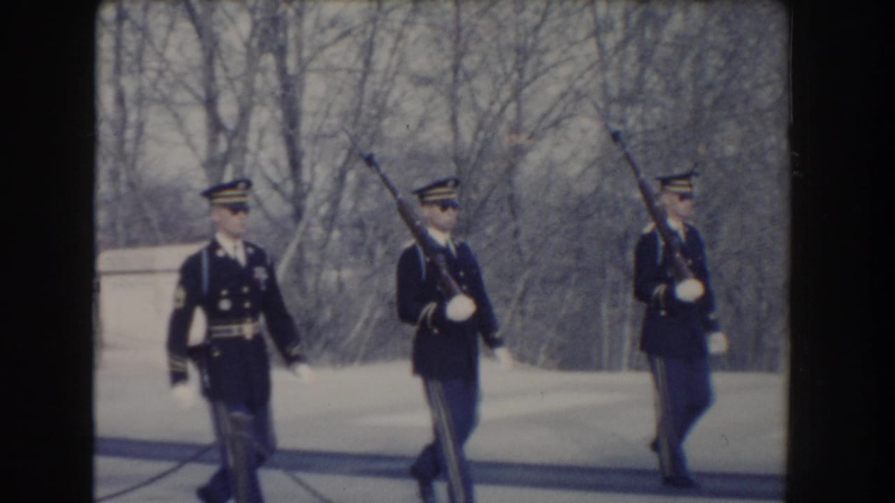 Three uniformed soldiers march with rifles near a pale stone structure and bare trees.